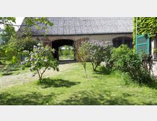 Garten mit grünem Rasen, Fliederbüschen und einem alten Gebäude im Hintergrund mit gewölbter Toröffnung und einer Fassade aus Natursteinen. Rechts sind grüne Fensterläden an einer Hauswand zu sehen, im Hintergrund stehen Tische und Stühle.