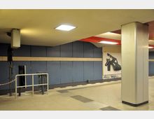 Unterirdischer Durchgang mit blauen Wandpaneelen, einer Reklametafel mit einem abgebildeten Schuh, sowie einer Metallabsperrung und Säulen. Die Decke ist gelblich, mit eingebauten Leuchtfeldern, und der Boden hat geflieste Muster.