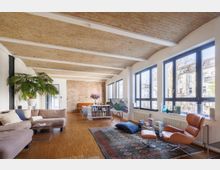 Wohnzimmer in einem Loft mit Ziegelsteindecke und Holzboden, großen Fenstern mit Blick auf eine Nachbarschaft und moderner Einrichtung wie einer braunen Ledersesselgruppe, einem Ecksofa, einem Teppich und Bücherregalen. Im Hintergrund ist eine Backsteinwand sichtbar, dekoriert mit Pflanzen und Kunstwerken.