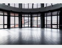 Moderne, lichtdurchflutete Innenraumansicht mit großen bodentiefen Fenstern und dunklen Metallrahmen, die einen Blick auf Außengebäude mit Glas- und Stahlkonstruktionen ermöglichen; der Boden besteht aus grauen Fliesen, die eine offene und minimalistische Atmosphäre schaffen.