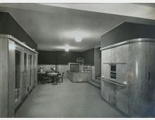 Schwarz-weiß-Fotografie eines Innenraums im Art-Déco-Stil. Zu sehen sind eine Kasse mit geschwungener Holzverkleidung rechts, ein Bereich mit Tischen und Stühlen links sowie eine kleine Verkaufstheke mit Glasvitrine im Hintergrund.