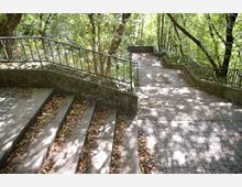 Treppenanlage in einem Wald mit Brüstungen aus Metall, flankiert von Bäumen. Der Boden der Wege besteht aus unregelmäßigen Pflastersteinen, und das Herbstlaub bedeckt teilweise die Stufen.