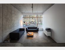 Wohnzimmer mit Betonwänden und moderner Einrichtung, bestehend aus zwei schwarzen Sofas, einem niedrigen Holztisch und einer weißen TV-Kommode mit Audiogeräten. Ein großes Fenster bietet Ausblick auf einen Wald mit Bäumen.