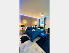 Modern eingerichtetes Schlafzimmer mit einem großen Bett, das mit blauer Bettwäsche und abgestimmten Kissen dekoriert ist. Helle Wände mit einer Holzvertäfelung im Kopfbereich, akzentuiert durch warmes Licht, hinterlegt mit indirekter LED-Beleuchtung; dazu passende Vorhänge, ein gepolsterter Hocker und Teppiche in Dunkelblau.