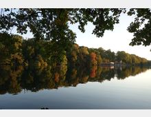 Ein See in ruhiger Umgebung, umgeben von herbstlich gefärbten Bäumen, die sich im Wasser spiegeln. Im Hintergrund befinden sich zwei gelbe Gebäude und ein Holzsteg am Ufer, teils verdeckt von den Baumkronen im Vordergrund.
