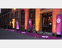 Backsteinfassade eines historischen Gebäudes mit großen Rundbogenfenstern, davor ein beleuchteter lila Teppich mit Absperrungen und Werbesäulen des Fernsehsenders Sat.1. Im Hintergrund ist ein Eingang mit Glastür sichtbar.