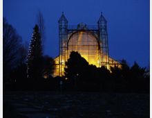 Ein beleuchtetes Glasgebäude, vermutlich ein Gewächshaus, bei Nacht. Im Vordergrund befinden sich dunkle Baum- und Strauchkonturen, während das Gebäude mit symmetrischen Türmen und einer großen Bogenscheibe im Zentrum gelb erleuchtet ist.