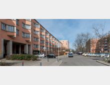 Wohnsiedlung mit mehreren vier- bis fünfstöckigen Mehrfamilienhäusern in geometrischem Stil mit orangefarbenen Fassaden und weißen Fenstern. Die Straße ist gesäumt von parkenden Autos, vereinzelten Bäumen und Gehwegen.