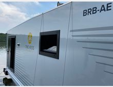 Ein modernes Hausboot mit der Aufschrift „The Kiss Klostersee“ und einem gelben Logo, das stilisierte Wellen und ein Boot zeigt. Im Hintergrund ist ein ruhiger See mit Wald am Ufer zu sehen, der Himmel ist teils bewölkt.