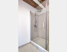 Moderne Dusche mit Glaswänden und einer Regendusche in einem hell gefliesten Badezimmer; die Decke zeigt freiliegende Backsteinbalken.