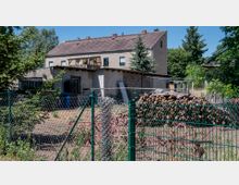 Ein ländliches Grundstück mit einem einfachen Wohngebäude aus Putz und Ziegeln, versehen mit einem Satteldach. Im Vordergrund befinden sich ein Maschendrahtzaun, Stapel von Holzscheiten und mehrere verstreute Gegenstände. Im Hintergrund sind Bäume und umliegende Gebäude zu sehen.