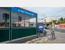 Außenansicht des U-Bahnhofs Yorckstraße in Berlin mit einem dunkelgrünen Eingangsbereich und einer Beschilderung. Im Hintergrund sind Wohngebäude, eine Straßenbrücke mit einer S-Bahn sowie Fahrräder und ein Bushaltestellenschild zu sehen.