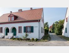 Eckhaus in einem Wohnviertel mit weißer Fassade, roten Fensterrahmen, grün lackierten Fensterläden und einem roten Ziegeldach. Vor dem Gebäude befindet sich eine gepflasterte Straße, die in eine schmale Gasse zwischen weiteren Häusern mit ähnlicher Architektur führt.