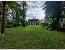 Ein großer Garten mit einer Rasenfläche, umgeben von Bäumen. Im Hintergrund befindet sich ein modernes zweistöckiges Wohnhaus mit einer Terrasse im Obergeschoss, daneben eine Rutsche und ein Klettergerüst für Kinder.