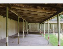 Ein offener Unterstand mit einem Dach aus Holzplanken, das von grünen Säulen gestützt wird. Der Boden besteht aus Beton, und an den Seiten befinden sich teils verwitterte, helle Wände; rechts ist eine Wiese zu sehen.