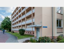 Wohngebäude mit mehreren Etagen und Balkonen in heller Farbe, umgeben von gepflegten Grünflächen und Sträuchern. Rechts neben dem Eingang befindet sich ein blaues Straßenschild mit der Aufschrift "Heinrich-Heine-Str. 41".