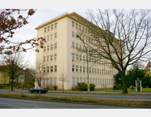Beigefarbenes, sechsstöckiges Bürogebäude mit zahlreichen Fenstern, umgeben von kahlen Bäumen und einer Straßenkreuzung im Vordergrund. Rechts im Bild befindet sich eine rote Telefonzelle auf einem Gehweg.