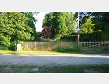 Ein ländlicher Weg mit einer niedrigen Holzlattenumzäunung, die eine Wiese mit hohen Bäumen und Buschwerk umgibt. Im Hintergrund sind Laubbäume und eine kleine Lichtung sichtbar.