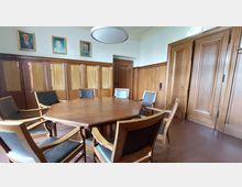 Holzgetäfelter Konferenzraum mit einem achteckigen Holztisch, umgeben von gepolsterten Stühlen mit Holzrahmen. An der Wand befinden sich Porträtgemälde, und die Türen bestehen ebenfalls aus Holz.