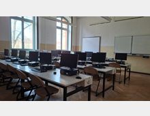 Computerraum/Klassenzimmer mit mehreren Reihen von Tischen, darauf Monitore, Tastaturen und Desktop-PCs, dazu Stühle. Große hohe Fenster lassen Tageslicht hinein; vorne an der Wand hängen Whiteboard und Tafel unter Deckenleuchten.