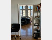 Blick durch eine offene Tür in ein Büro mit Parkettboden, einem schwarzen Ledersessel und einem großen Fenster, durch das ein Gebäude und blauer Himmel sichtbar sind. Im Vordergrund ein Drehstuhl und ein Regal mit Akten und Büchern an der rechten Wand.