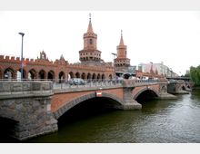 Historische Backsteinbrücke über einen Fluss mit zwei Türmen im neugotischen Stil, Querbögen und einer Kombination aus Fahrbahn und Fußgängerbereich. Im Hintergrund moderne Gebäude und Bäume am Flussufer.