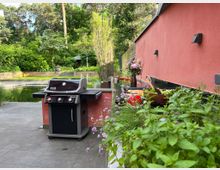 Terrasse eines modernen Wohnhauses mit roter Fassade und grauen Steinplatten, daneben ein Gasgrill und eine Abstellfläche mit Pflanzen. Im Hintergrund liegt ein Naturpool und ein dicht bewachsener Garten mit Bäumen und Sträuchern.