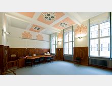 Geräumiges Besprechungszimmer mit hohen Decken, verziertem Stuck in Rosa und Weiß, großen Holzvertäfelungen an den Wänden und bodentiefen Fenstern, die viel Tageslicht hereinlassen. Ein langer Konferenztisch mit schwarzen Stühlen dominiert den Raum, der zudem durch seitliche Heizkörper und moderne Hängelampen ergänzt wird.