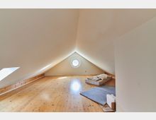 Minimalistisch eingerichteter Dachbodenraum mit schrägem Giebeldach, hellem Holzfußboden, einem runden Fenster an der Stirnseite und einer schrägen Dachluke links. Auf dem Boden liegen eine Matratze mit Kissen sowie eine graue Decke.