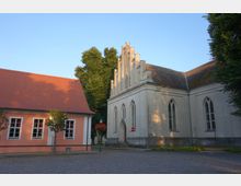 Historisches Gebäudeensemble mit einer kleinen Kirche in gotischem Stil auf der rechten Seite, gekennzeichnet durch spitzbogige Fenster und eine gestufte Giebelfassade, daneben ein rosa Gebäude mit roten Dachziegeln und weißen Fenstereinfassungen, umgeben von Bäumen und einer gepflasterten Straße.