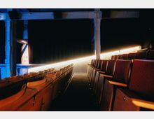 Innenansicht eines kleinen Theaters oder Kinosaals mit roten Polstersitzen in Reihen angeordnet. Im Hintergrund ist ein schwarzer Vorhang und eine schräg verlaufende Lichtlinie zu sehen, beleuchtet von warmem Licht.