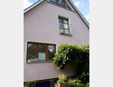 Ein Einfamilienhaus mit rosafarbenem Putz und einem Satteldach. Holzrahmenfenster prägen die Fassade, eines davon hat ein schmiedeeisernes Geländer im oberen Stockwerk. Ein Rankgewächs bedeckt einen Teil der Hauswand, und im Vordergrund stehen Topfpflanzen.