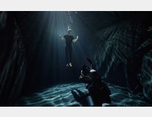 Underwater cavern-like space with dark rock walls and a flat floor, lit by sunbeams and rippling light patterns from the surface above. A scuba diver with equipment is near the bottom, with floating particles visible in the water.