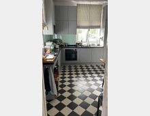 Moderne Küche mit grauen Einbauschränken, schwarzer Arbeitsplatte und einem Backofen. Der Boden ist mit schwarz-weiß karierten Fliesen gestaltet, und ein Fenster mit gestreifter Jalousie lässt Tageslicht herein.