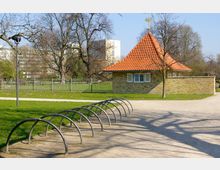 Kleines Gebäude mit rotem Ziegeldach und geschlossenen Fensterläden in einem Parkgelände, umgeben von Bäumen und einem Zaun. Im Vordergrund befinden sich geschwungene Fahrradständer auf einem unbefestigten Weg.