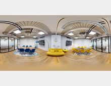 Modernes Konferenzraum-Interieur mit zwei Bereichen, die durch eine gelbe Couch getrennt sind. Linker Bereich mit blauem Tisch und blauen Stühlen, rechter Bereich mit ovalem Tisch und gelben Stühlen, dazu Wände mit Glaselementen, weißen Tafeln und TV-Bildschirmen; sichtbare Deckenrohre und Holzboden.