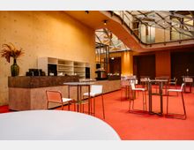 Innenansicht eines modernen Cafés mit roten Teppichböden, hellen Wänden und minimalistischen Möbeln. Es gibt hohe Tische mit weißen Stühlen sowie eine Theke mit Regalen voller Gläser und einer Kaffeemaschine, umgeben von einer Glas- und Stahldeckenkonstruktion.