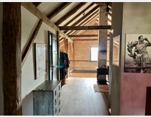 Dachbodenraum mit freiliegenden Holzträgern und einer ziegelsteinernen Wand im Hintergrund. Im Vordergrund befinden sich eine Kommode und Bilder an den Wänden, rechts ein Schwarz-Weiß-Foto und ein kleiner Schreibtisch mit Stuhl.