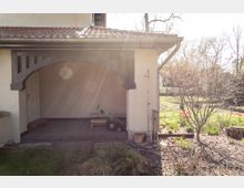 Außenansicht eines überdachten Eingangsbereichs mit einem Rundbogen aus Holz, der an ein Gebäude mit hellem Putz grenzt. Im Vordergrund befindet sich ein Garten mit blühenden Bäumen und Beeten, die teils von Sonnenlicht erhellt werden.