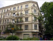 Eckgebäude in einem Wohnviertel mit historischer Fassade im Stil der Gründerzeit, verziert mit Stuck und verzierten Gesimsen. Im Erdgeschoss befinden sich Geschäfte, während die oberen Etagen Balkone mit Pflanzen haben; umgeben von Bäumen und Straßenlaternen.