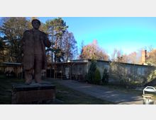 Steinfigur eines Mannes auf einem Sockel in einem Außenbereich mit Bäumen im Hintergrund. Neben der Statue steht ein flaches Gebäude mit Gitterfenstern, das von einem gepflasterten Weg und etwas Grünfläche umgeben ist.