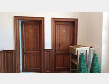 Zwei Holztüren mit den Buchstaben „H” und „D” auf heller Wand, umgeben von holzvertäfelten Wandelementen, daneben ein kleiner Tisch und künstliche grüne Pflanzen.