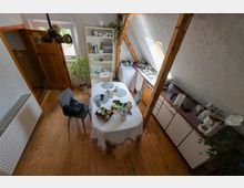 Kleine Dachgeschossküche mit Holzfußboden, sichtbaren Holzbalken und schrägen Wänden; in der Mitte steht ein ovaler Esstisch mit Tischdecke und Stühlen. Entlang der Wand verläuft eine Küchenzeile mit Spüle unter einem Dachfenster, daneben ein offenes Regal mit Geschirr und ein Fenster zur Seite.