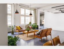 Heller, modern eingerichteter Raum mit einer Sitzecke aus Holzmöbeln, bunten Kissen und einem Holztisch auf einem beigefarbenen Teppich. Große Fenster lassen Tageslicht herein, umgeben von Pflanzen, und ein weißer Hintergrund im hinteren Bereich deutet auf ein Studio-Setup hin.