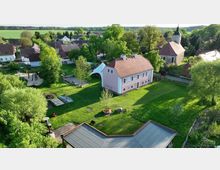 Luftaufnahme eines Dorfes mit einem großen Rasenbereich, auf dem ein zweistöckiges, rosa Gebäude mit rotem Ziegeldach steht. Im Hintergrund befinden sich eine Kirche mit Turm, weitere Häuser und Bäume, umgeben von Feldern und Wäldern.