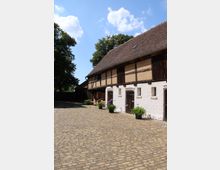Fachwerkgebäude mit weiß verputztem Erdgeschoss und dunkelbraunen Holzdetails, umgeben von einem gepflasterten Hof. Im Hintergrund sind Bäume und ein blauer Himmel mit wenigen Wolken zu sehen.
