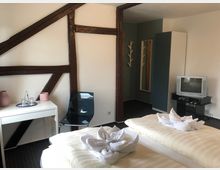 Ein schlicht eingerichtetes Hotelzimmer mit einem Doppelbett, dunklen Holzbalken an der weißen Wand, einer Sitzecke mit Tisch und Stuhl sowie einem kleinen Fernseher auf einem schwarzen Tisch. Im Hintergrund befinden sich ein Kleiderschrank, ein Wandspiegel und eine Garderobe.