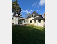 Rückansicht eines hell verputzten Einfamilienhauses mit dunklem Walmdach und großen Sprossenfenstern, teilweise als verglaster Anbau ausgeführt. Davor liegt eine gepflasterte Terrasse am Rasen, umgeben von Bäumen unter blauem Himmel.