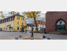 Ein gepflasterter Platz mit einer modernen Metallskulptur in der Mitte, umgeben von Herbstlaub, einem gelben Gebäude mit der Aufschrift „Jägerhof“ auf der linken Seite und einer roten Backsteinkirche mit einem großen Eingangstor auf der rechten Seite. Im Hintergrund sind weitere Gebäude und ein Baum zu sehen.