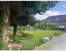 Garten mit dichtem Grün, einem großen Baum im Vordergrund, einer Rutsche und einem Spielbereich im Hintergrund. Im hinteren Bereich stehen ein Haus mit Schrägdach und eine Hecke, die den Garten umgeben; Wäsche hängt auf einer Leine zum Trocknen.